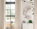 A styled interior room scene with a skeleton prop seated in a light wood rocking chair, cream upholstered chairs, beige curtains, a white cabinet, wall art, and black bat decorations.